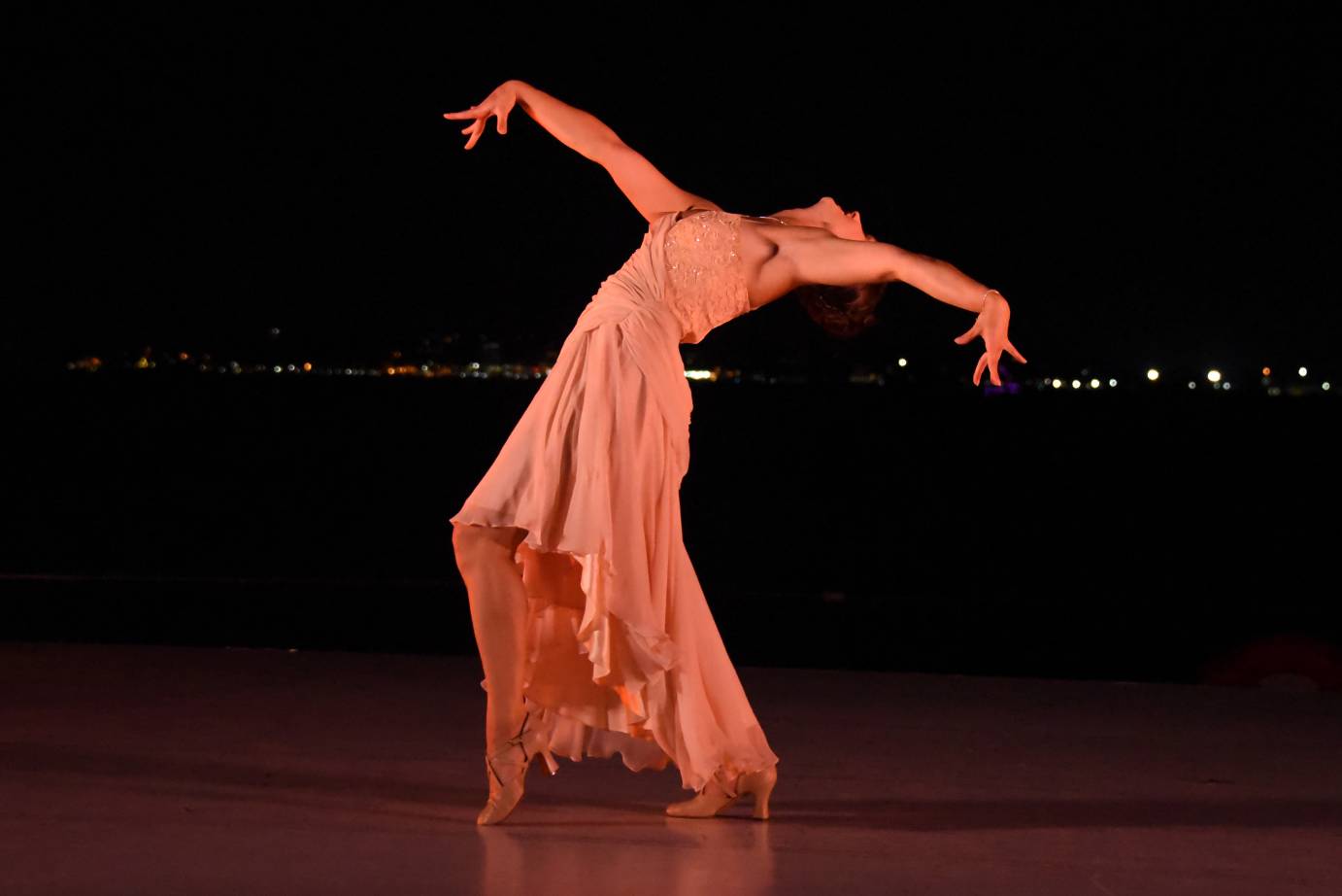 Jillian Linkowski stands in profile, wearing a long, side-cut peachy-pink gown. She is in a deep backbend with one heel popped. Her arms are extended like wings with stylized fingers.