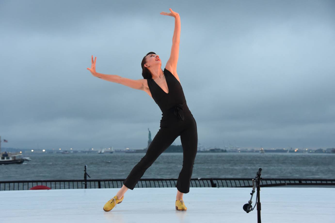 In a  black jumpsuit with a deep V-neck and yellow tap shoes, Demi Remick points one foot to the  side as her arms are lifted in a V, wrists flexed.