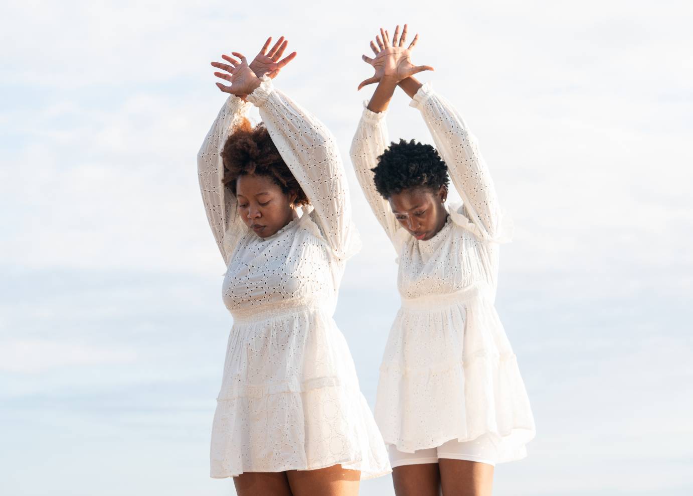Two Black women wear long-sleeved, white lace dresses. Kate Louissaint is to Nhyira Oforiwaa Asante's left. While looking down, their arms are lifted above their head, wrists crossed