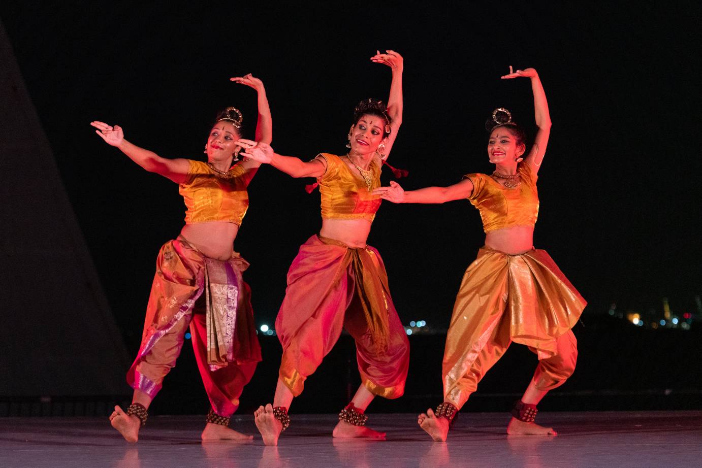 Three women in gold tops with short sleeves, blousy pants, and bells on their ankles are in a line. They lift one arm overhead with the palm flipped up as they extend one foot and the other arm toward the audience.