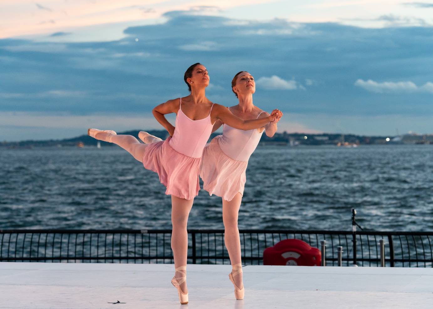 In pink leotards and chiffon skirts, Amanda Treiber and Giulia Farina stand in arabesque en pointe. Treiber is slightly behind Farina and holds her hand.