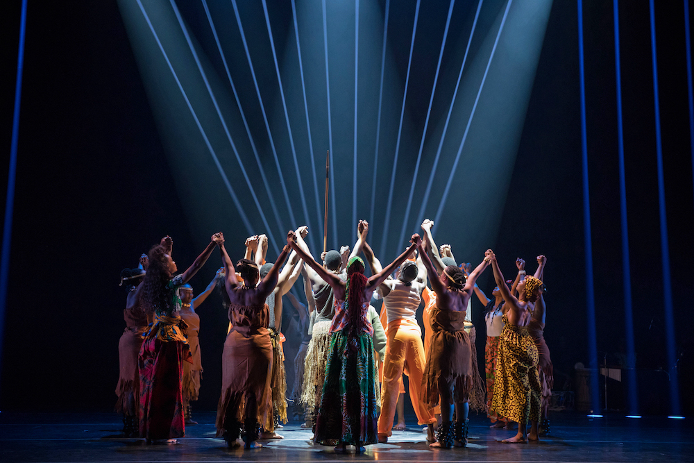 A clump of colorfully-clad dancers reach their hands to the sky. They stand in dark light with pinstripes of white light rising vertically