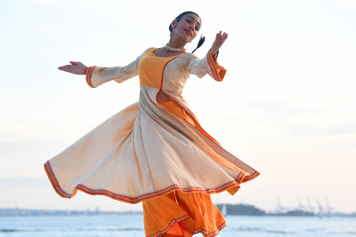 Parul Shah wears a flowing white dress with a yellow bodice and cuffs. With a smile on her face, she poses in profile, one hand extended backward, the other gently lifted.