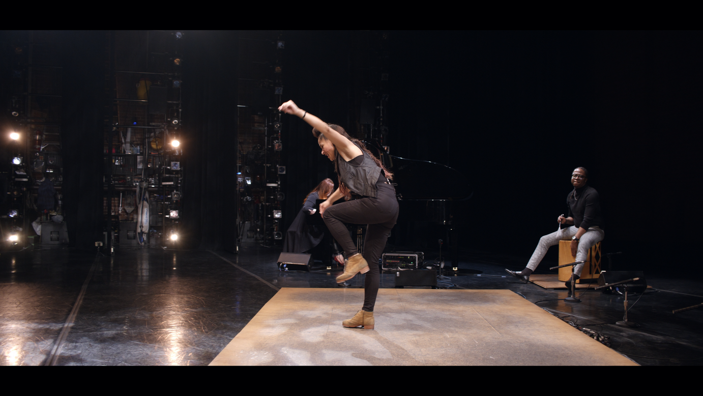 Ayodele Casel lifts one arm over her head. She is dressed in black.  A few musicians are situated around the wooden platform. 