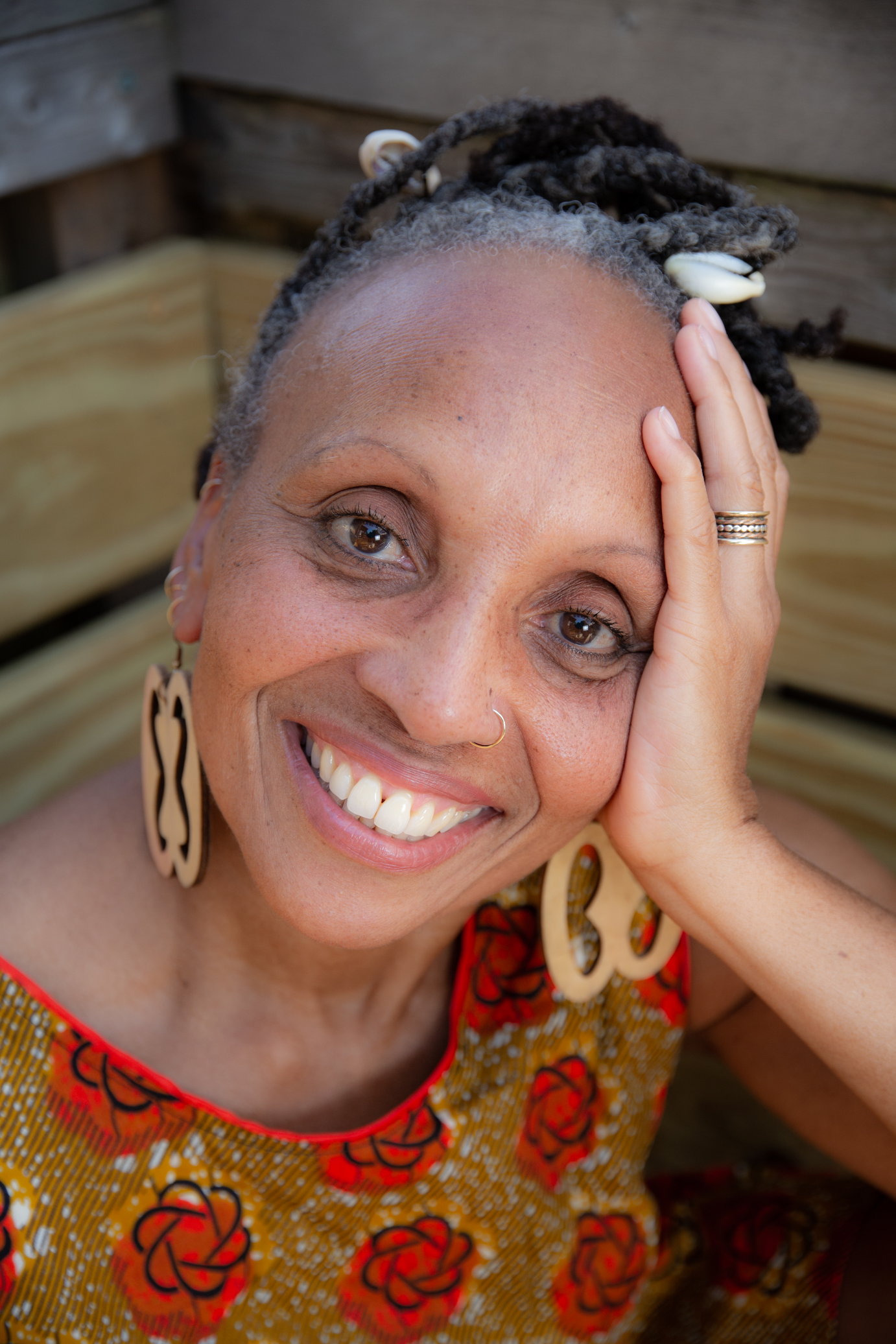 A headshot of Charmaine Warren in a yellow dress with red flowers. She is smiling and has one hand on her head.