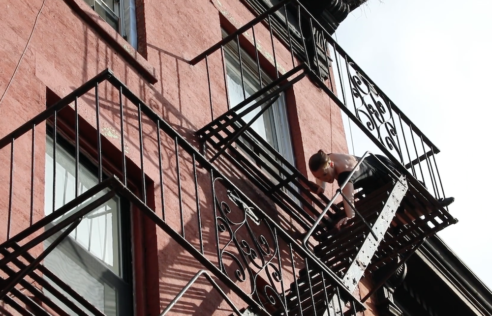 fairskinned woman in a black dress, seated on her black wrought iron fire escape lowering the upper half of her body on the descending stairs... building is red brick