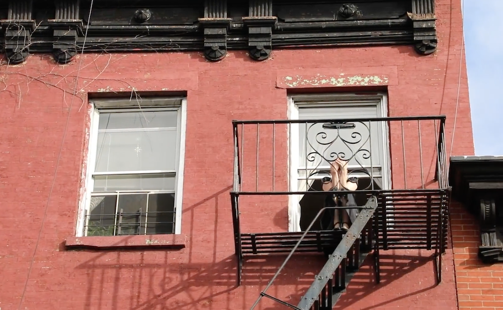 red Brick apartment building, black wrought iron fire escape, fair skinned woman dressed in black clutching her head in both hands as she sits on her window sill