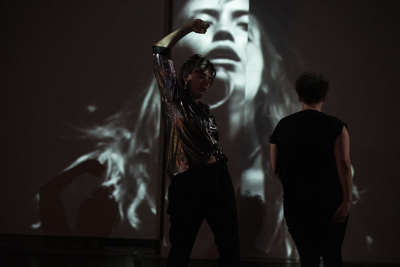 One dancer lifts an arm, casting a shadow against a black and white film of a woman with her mouth open. Beside her, a woman in a black top faces upstage.