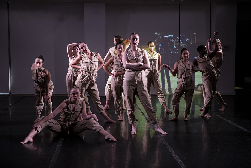 A group of women in beige jumpsuits gesture defiantly with crossed arms or hands on hips. They look unimpressed.