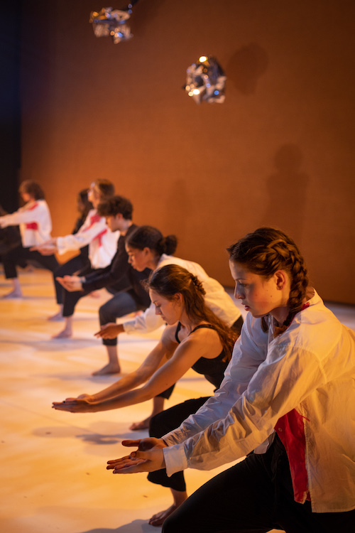a long line of teen dancers, all leaning on a bent leg with arms extended and hands folded together.