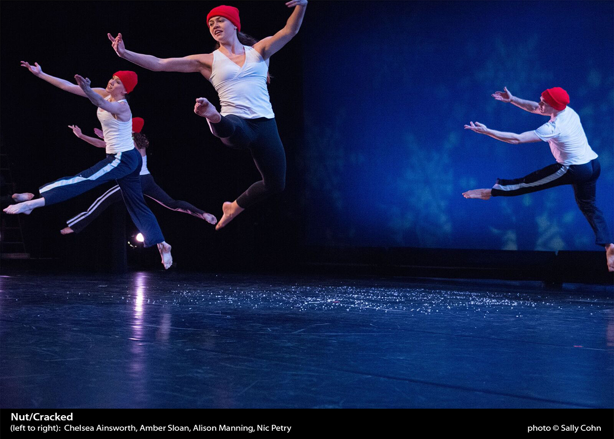 Four dancers in white tops, black sweatpants, and red beanies grand jeté with a bent back leg.