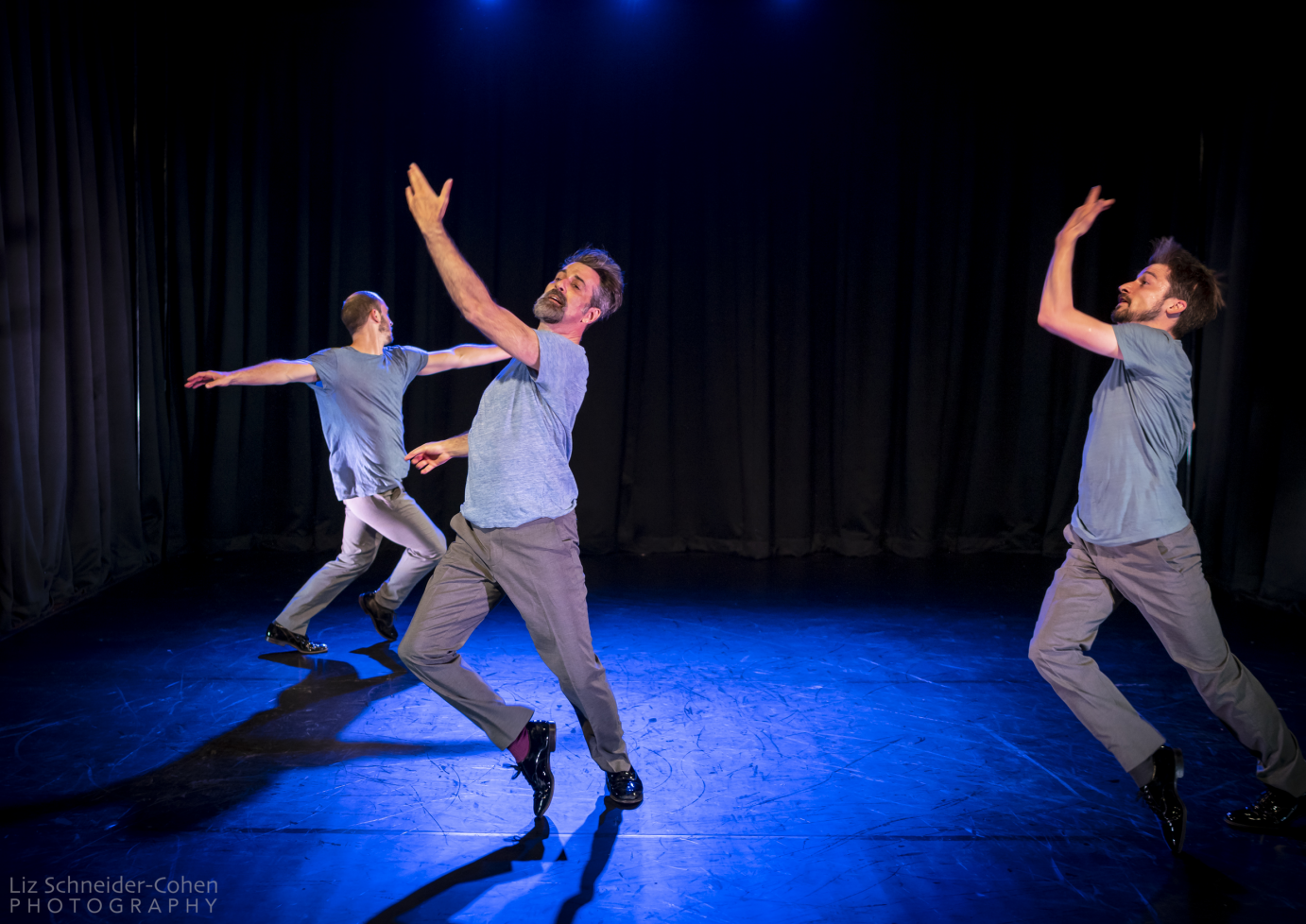 Three men in blue T-shirts and gray pants are arranged in a triangle. They are stepping forward with one foot and lifting one bent elbow in front of them. 