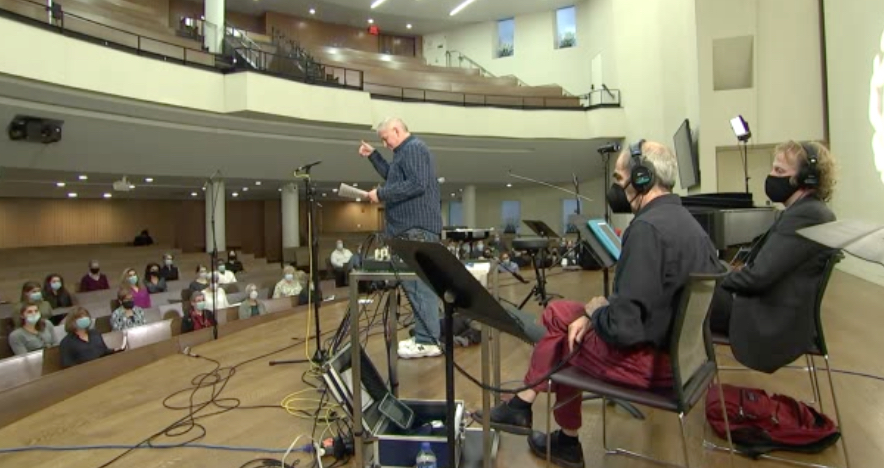 Men in headsets sit on a stage while a few scattered people sit in the audience.