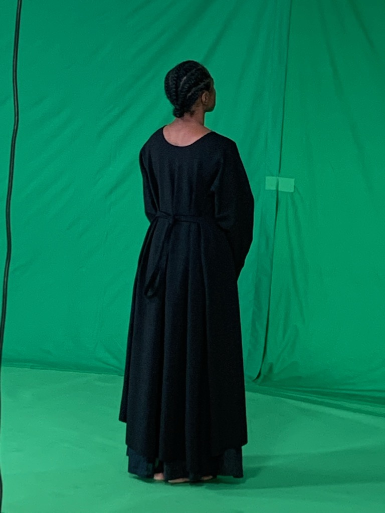 A black woman in a long black dress stands with her back to the camera. A green background frames her.
