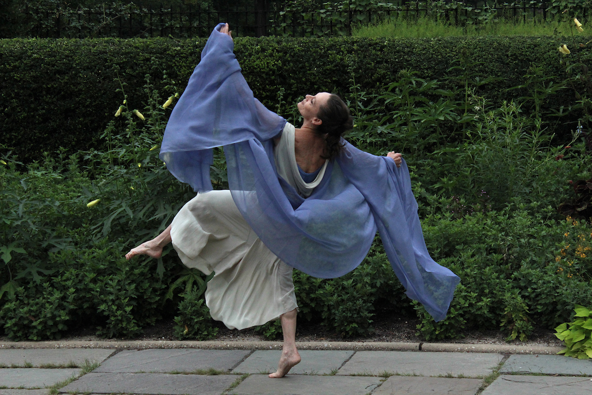 In a flowing white dress and periwinkle blue scarf, Christine Dakin faces away from the camera. She lifts her leg in a side attitude and makes a gentle arc with her arms.