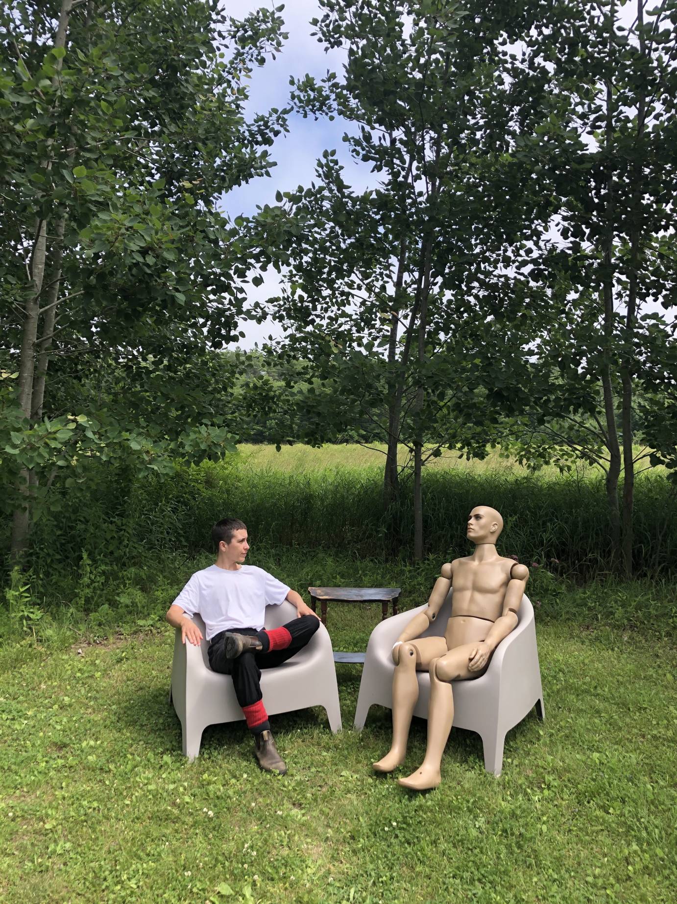 In a grassy glade, devynn emory and manny the mannequin sit in lawn chairs staring at each other