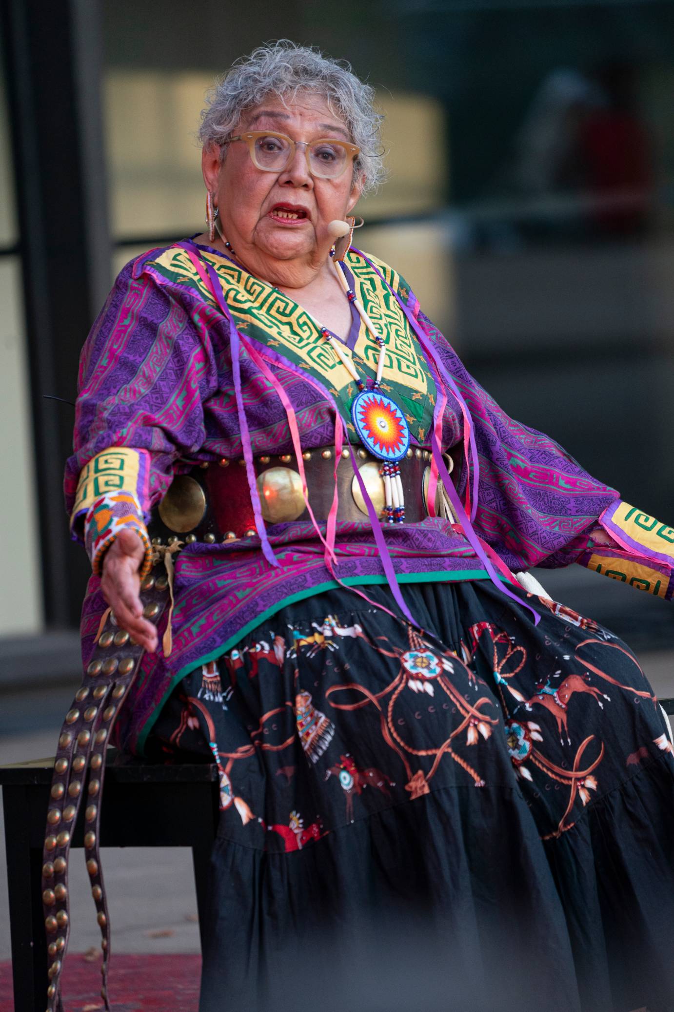 Muriel Miguel aka Bright Sun sits. She wears a purple tunic with graphic yellow accents on the wrists and neckline. She also has on Native American jewelry