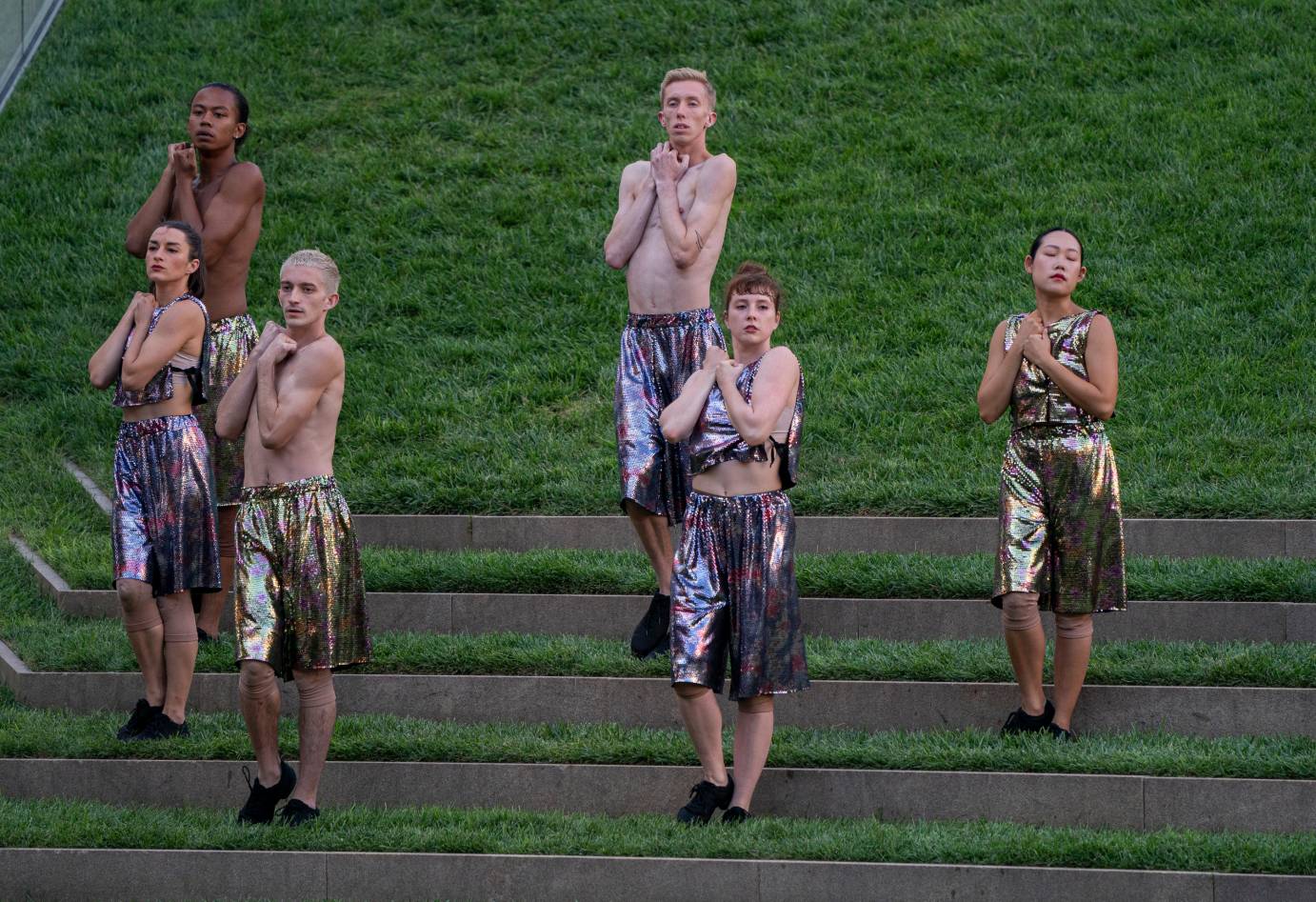 Against a grassy mound, dancers in metallic fabrics clutch their hands to their chests