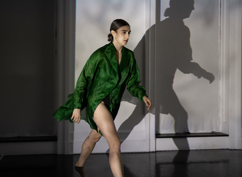 Wearing a green shirt dress, Xenia Mansour walks in profile. Her shadow looms ominously on  he white wall behind her.