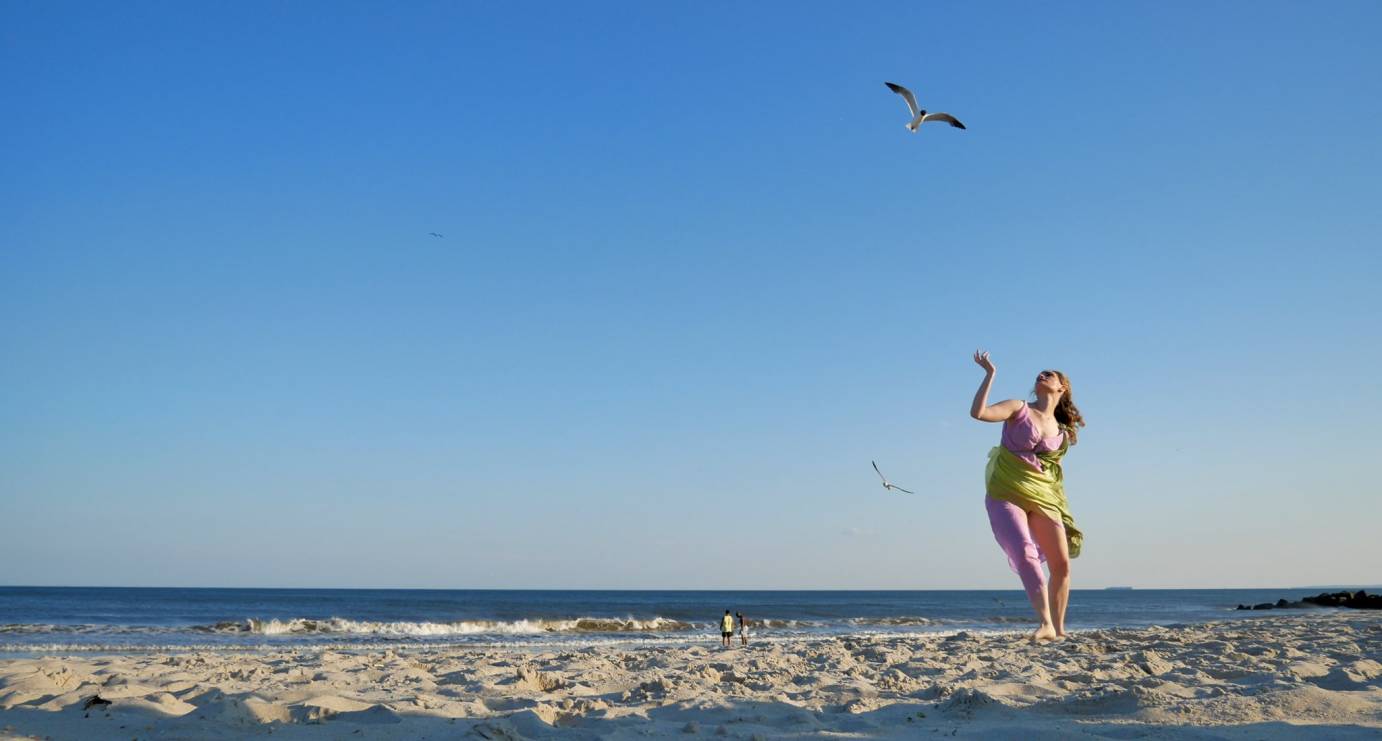 A woman in a lavender and green tunic poses on a beach with her arm up like she is holding a tray. Her body is tilted back. Two men by the water's edge are by the water's edge.