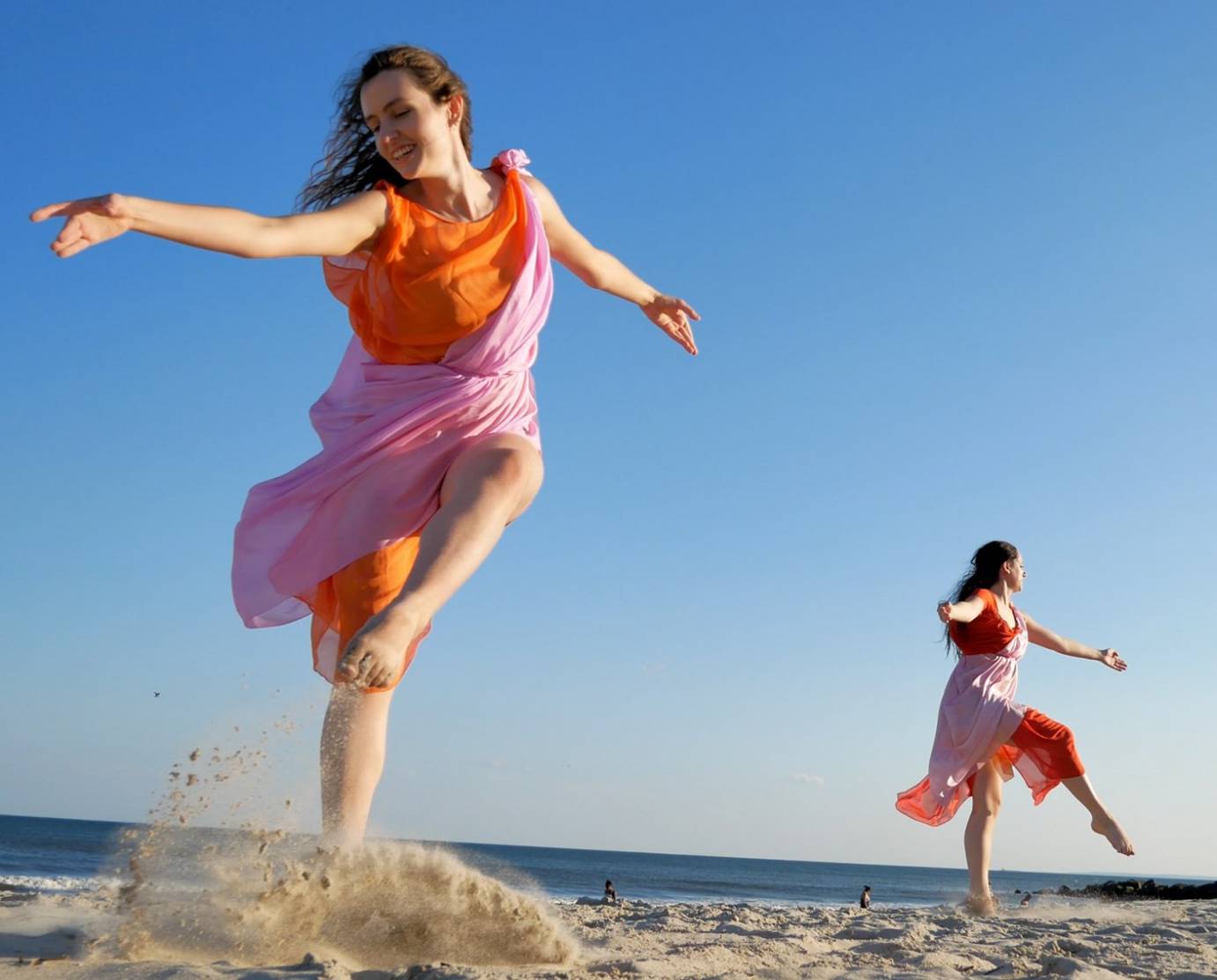 Against a blue sky, two women in orange and pink Grecian tunics lift opposite legs in attitude while smiling. 