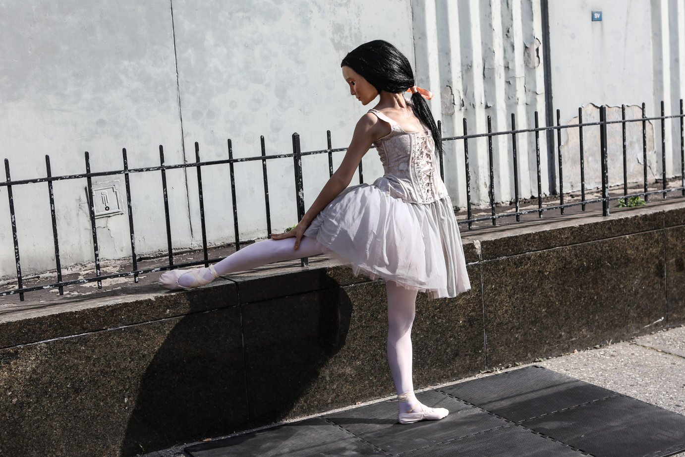A dancer with a plastic mask and wig evokes Edgar Degas' Little Dancer. She lifts a ballet-slippered foot to rest against a wrought-iron fence.