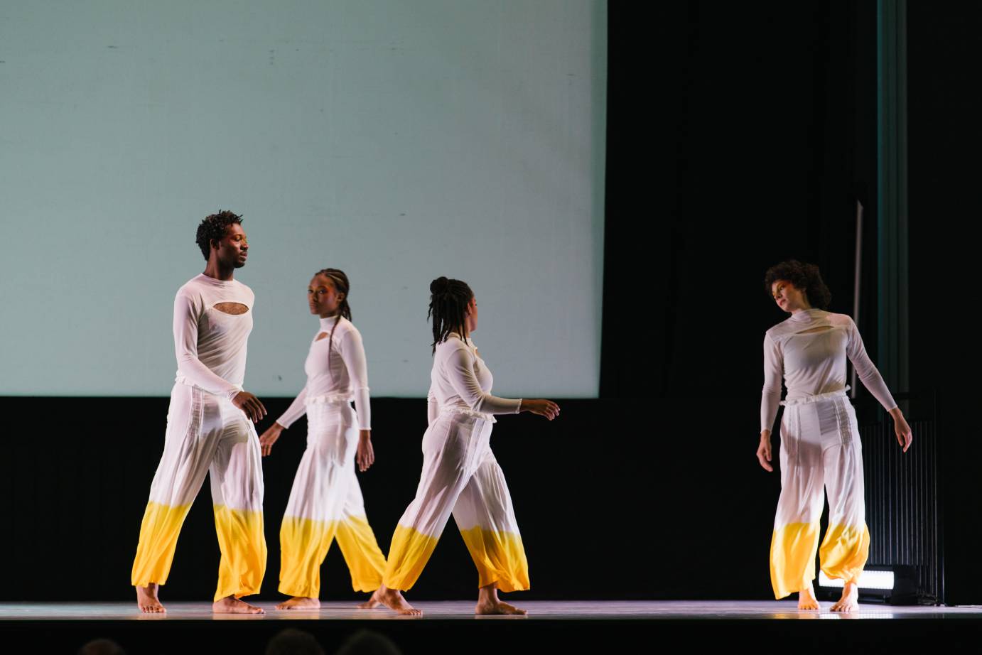 A quartet of dancers walk casually in a circle; one female dancer looks over her shoulder