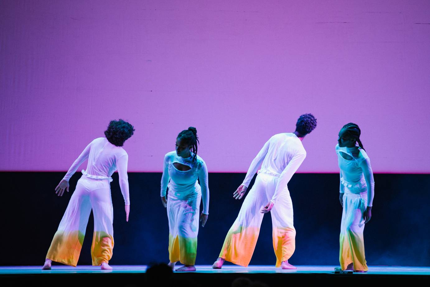 Two dancers lean toward stage left while two lean forward; they are all against a lilac-colored cyclorama