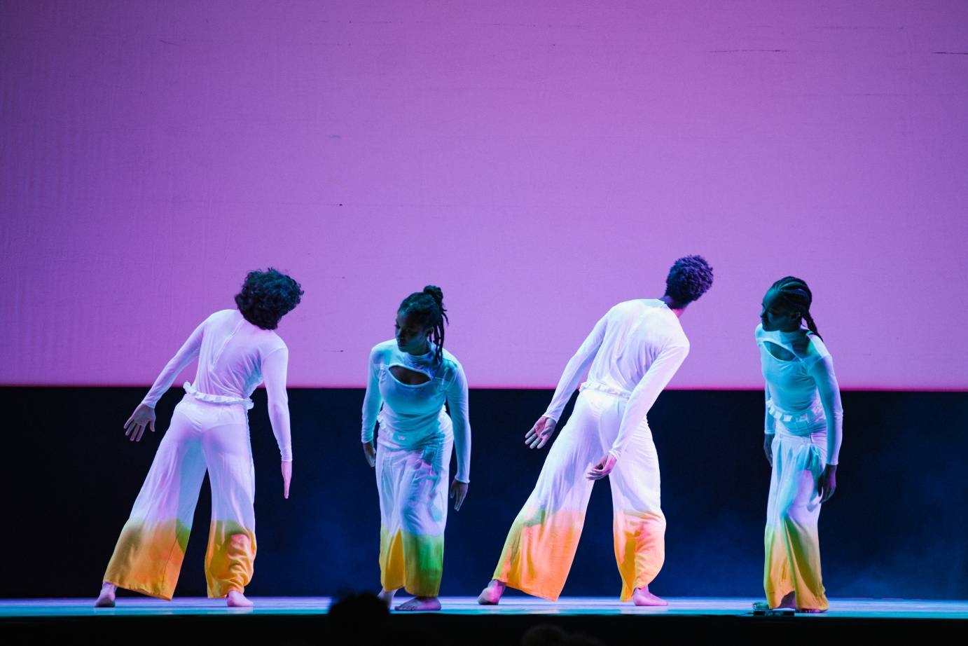 Two dancers lean toward stage left while two lean forward; they are all against a lilac-colored cyclorama 