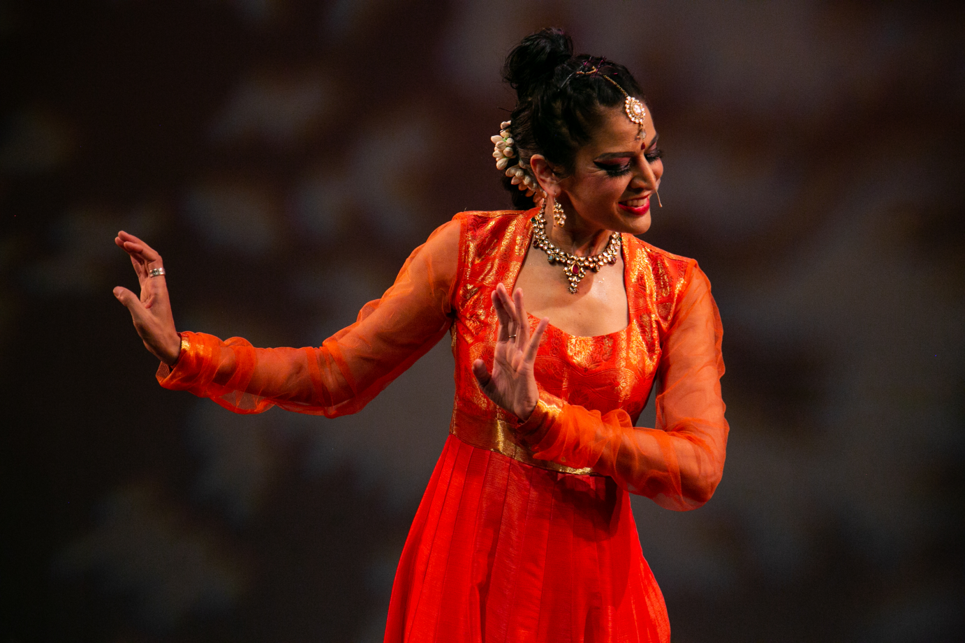 Rachna Nivas wears a bright red sari. She has both arms gently curved to the left. She is smiling.