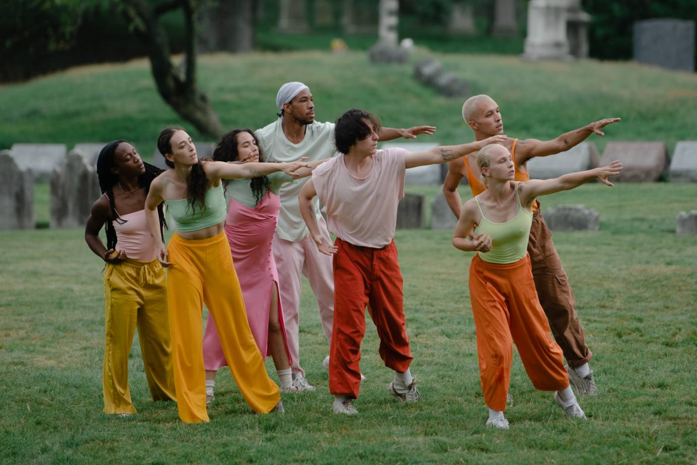 Seven dancers stand in a clump with one arm extended, the other bent at their sides. 