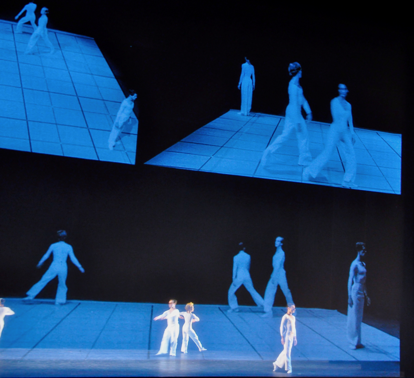 The film is split into two panels. In front and on stage, four dancers walk.