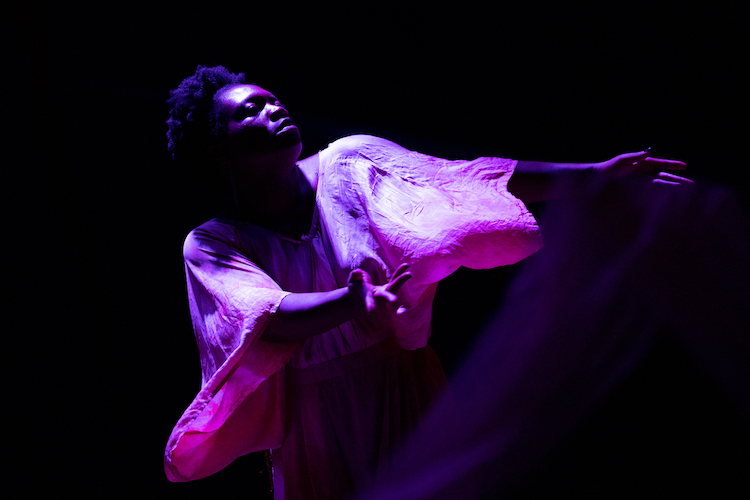 black woman against a black background wearing pink silky dress the light shines on her forehead and cheeks. 