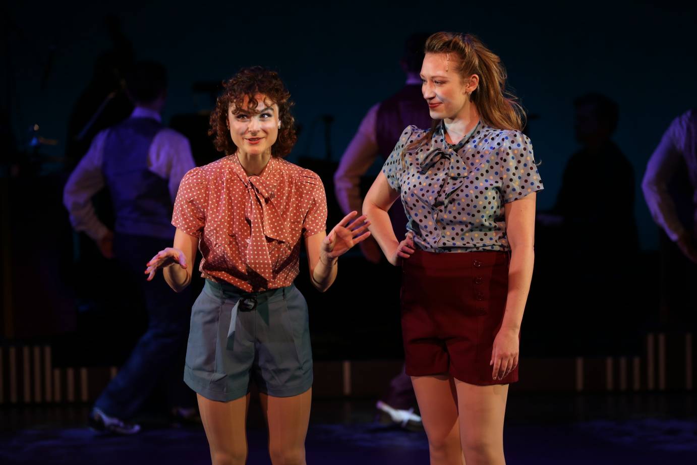 In a red-print blouse and denim shorts, Melanie Moore leans forward smirking, as if she'd telling a story to the blonde woman standing next to her.