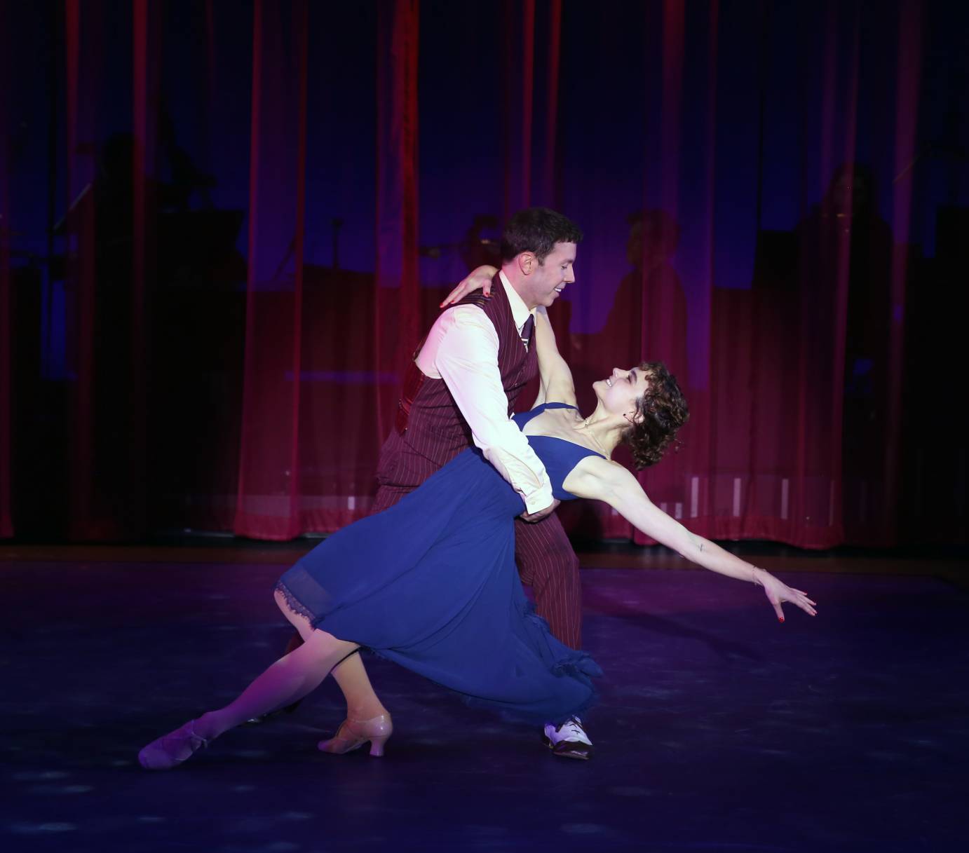 A man in a vest dips Melanie Moore as she creates a diagonal line from her extended leg to one arm lifted. She wears a low,cut, circle-skirt blue dress.