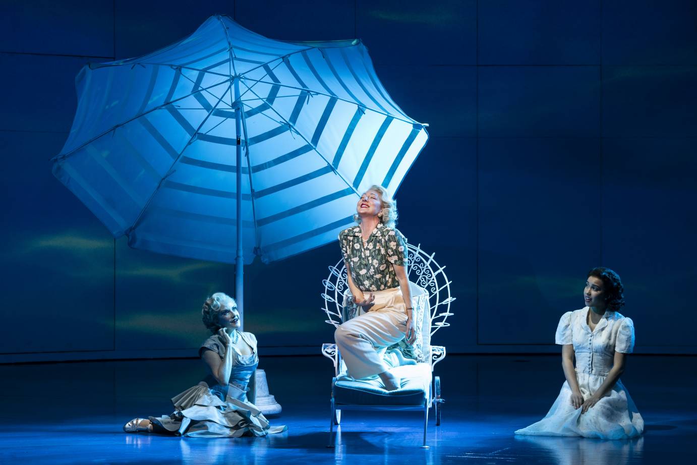 In a Hawaiian shirt, Carmen Cusack kneels on a white chair under a large umbrella with two women flanking her. The two women sit with their legs under them.