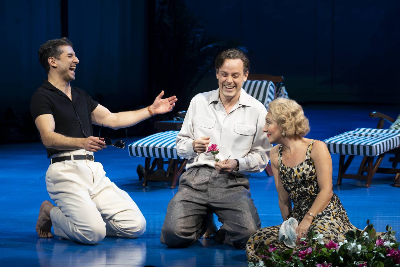 The actors playing Cary Grant, Aldous Huxley, and Clare Booth Luce kneel in a clump, laughing and gesticulating.