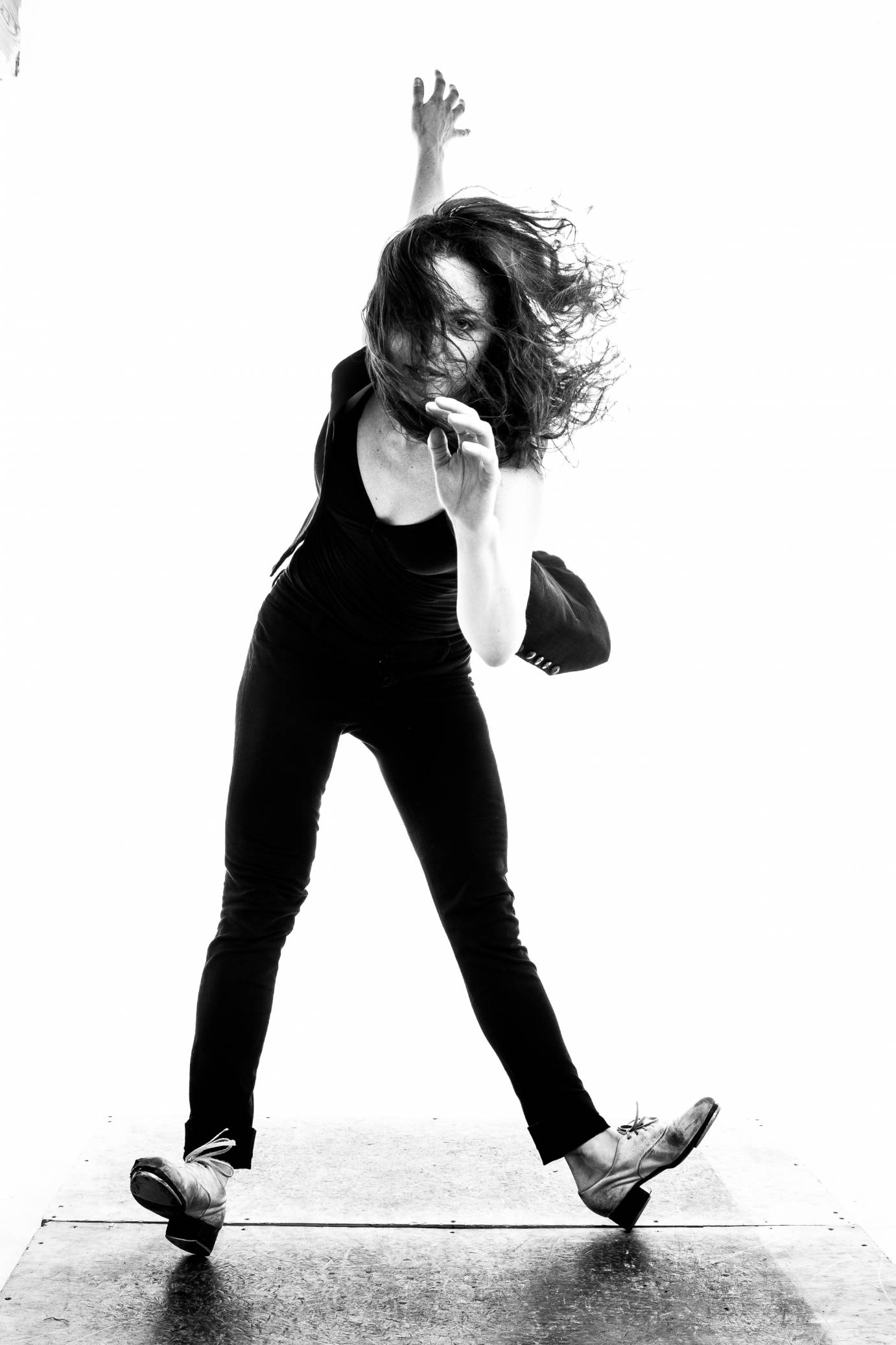 in a black and white photo, Michelle Dorrance stands on her heels in tap shoes, her hair flying in her face.
