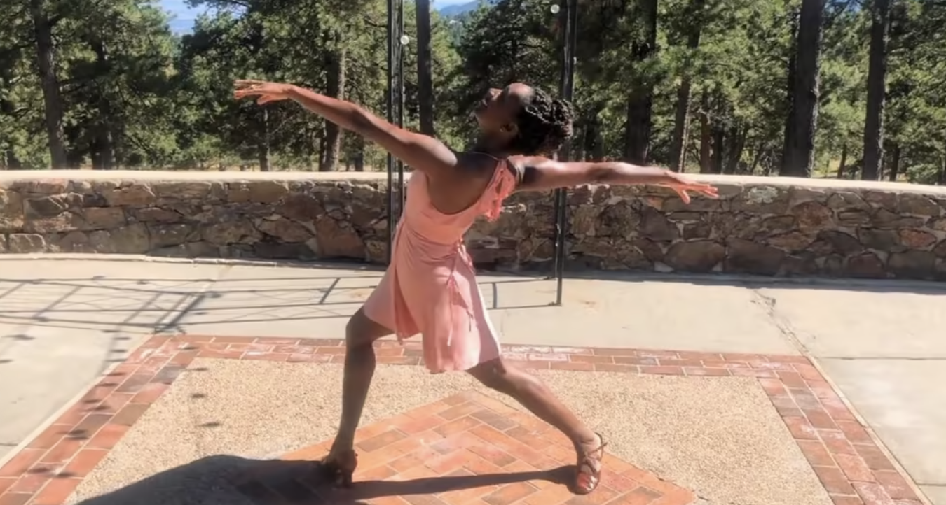 A woman in a peach dress extends her arms and legs in a gliding lunge. She is outdoors with trees behind her.