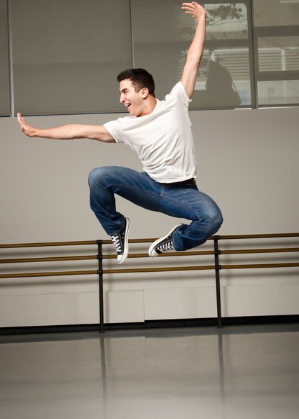 Jess LeProtto jumps in the air with both  legs tucked underneath him and his arms extended—one up and one to the side, both with flexed wrists. He wears blue jeans, a white T-Shirt, and Converse sneakers.