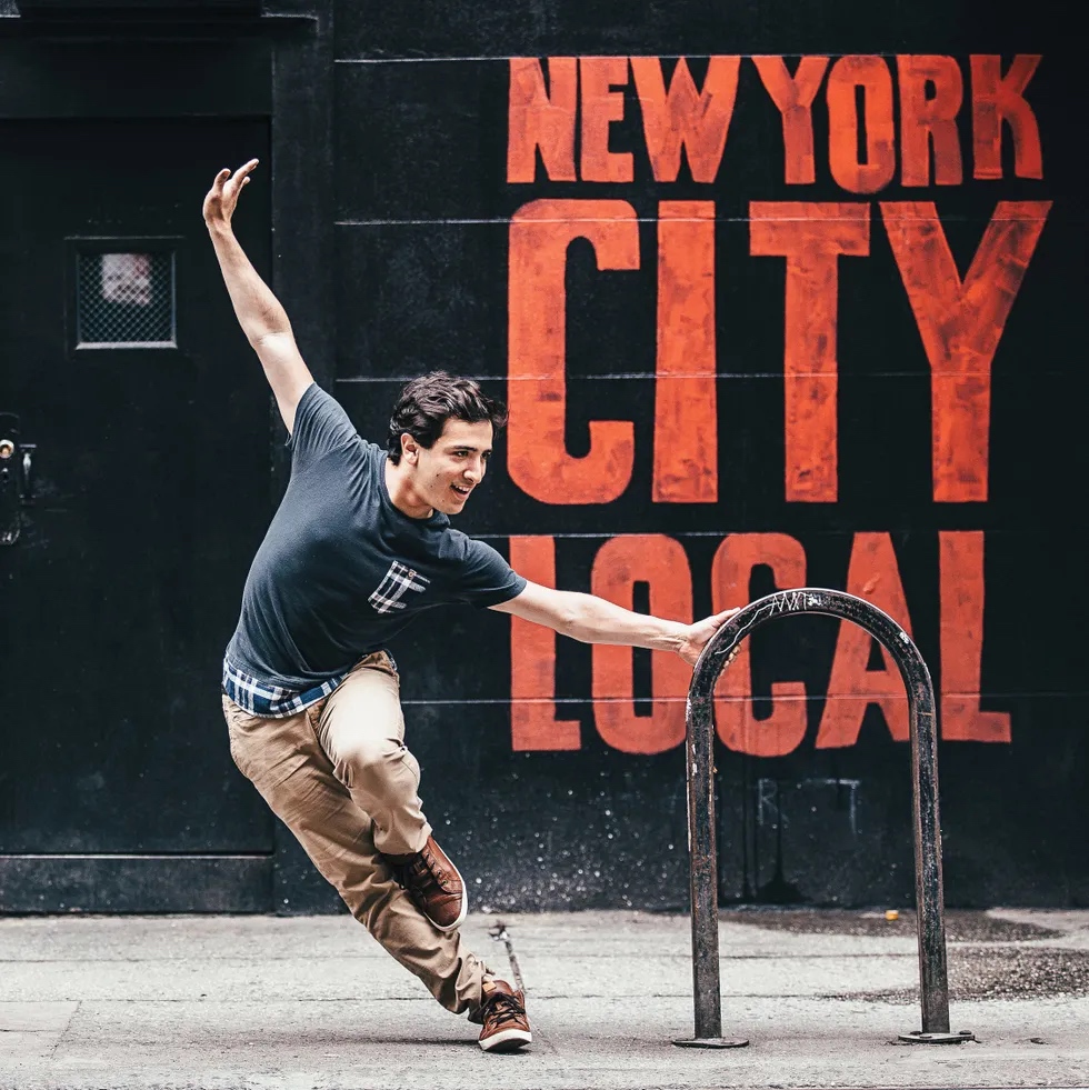 A man holds on to a U-shaped pole. He pulls his body away from the pole and lifts  one leg so that the foot is against his ankle. New York Local is written on the photo in red.