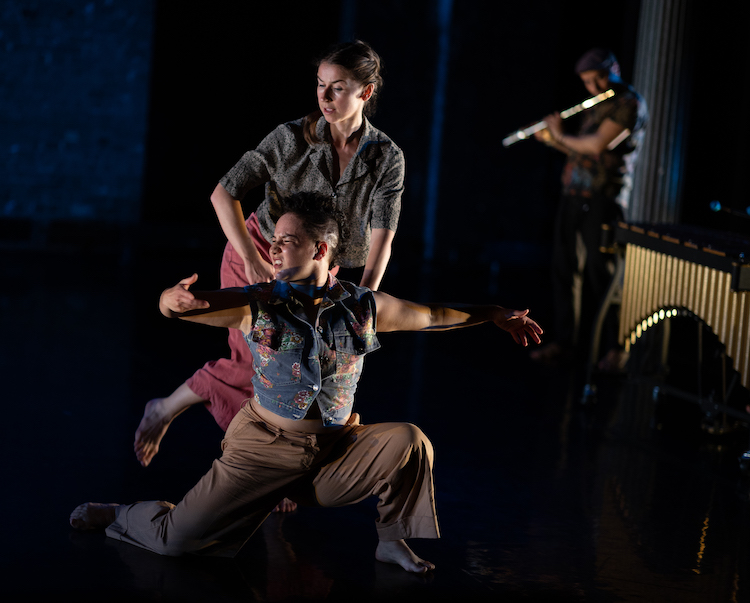 a duet with two women one in a semi squat on the floor with a grimace on her face, above her another dancer appears to listen and watch with concern, a flutist plays in the background