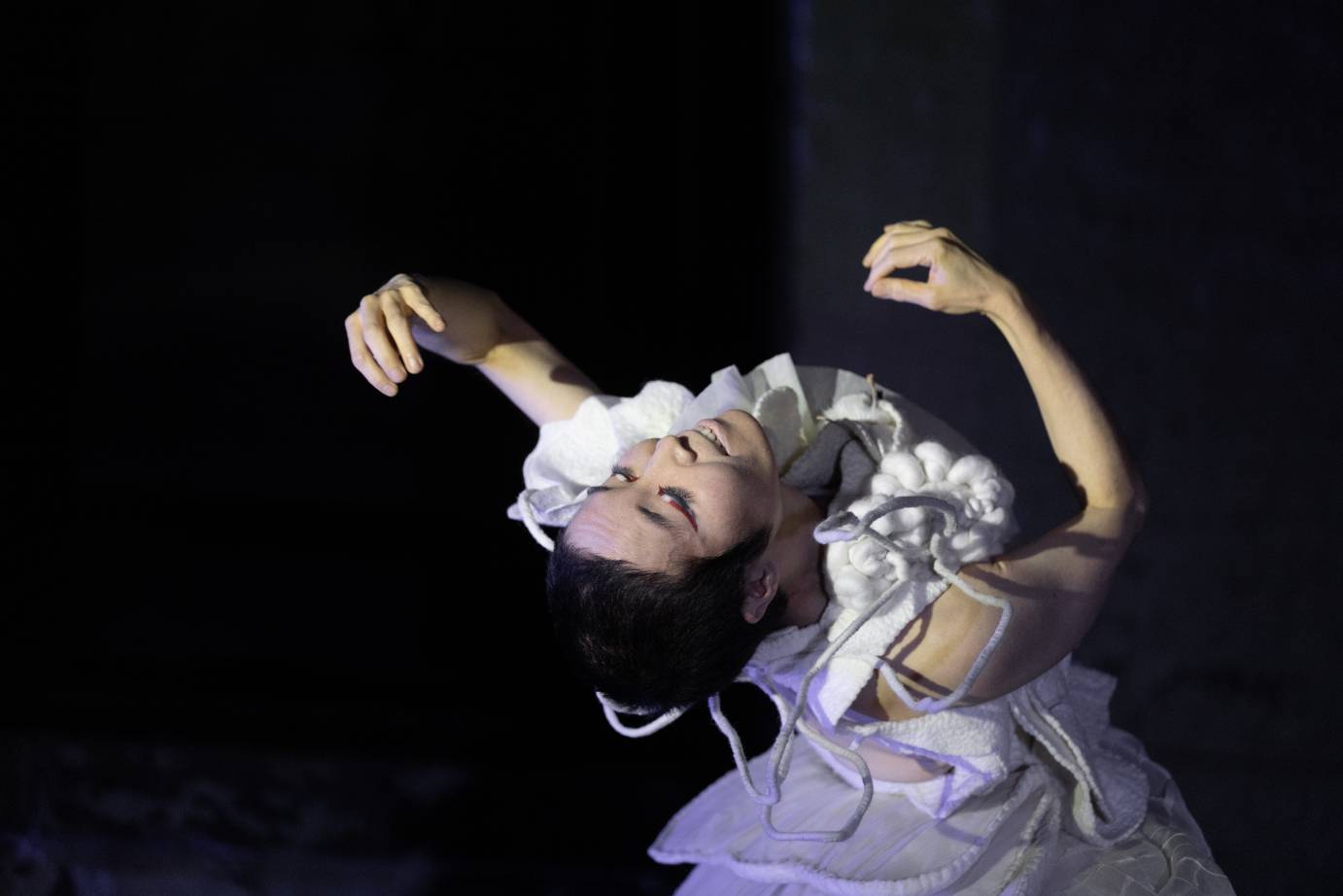 A woman in a ruffly white dress bends backward to the camera, her fingers crimped