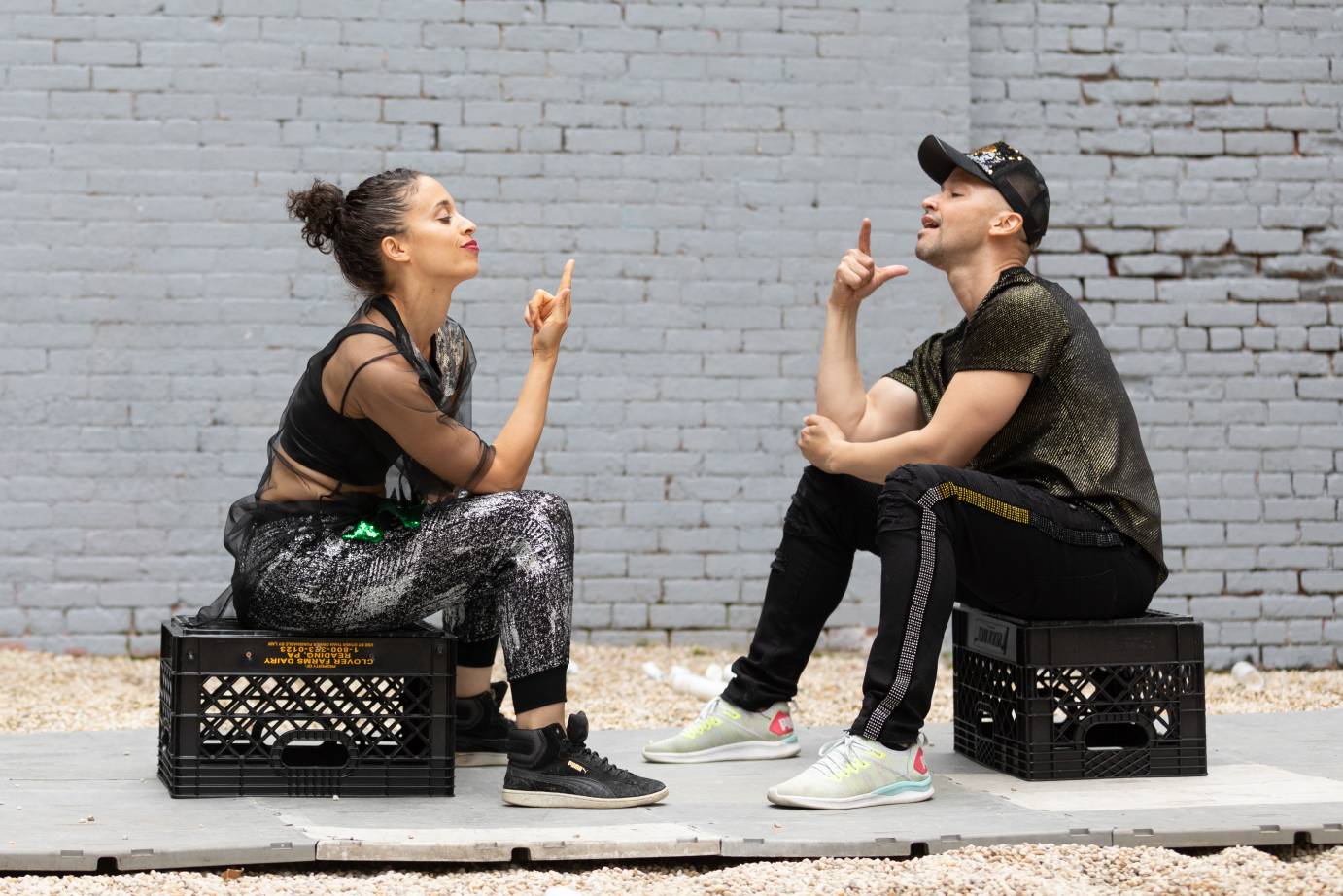 A man and a woman sit on black milk crates; both point a finger in the air