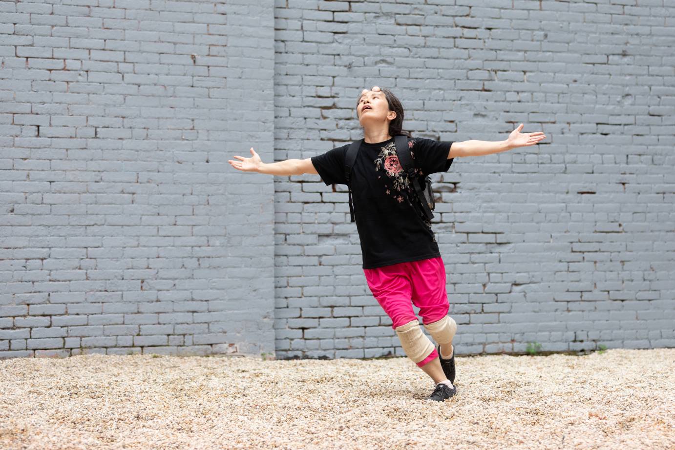 A woman in a black  t-shirt ad hot pink pants opens her arms wide and looks upward with her mouth open