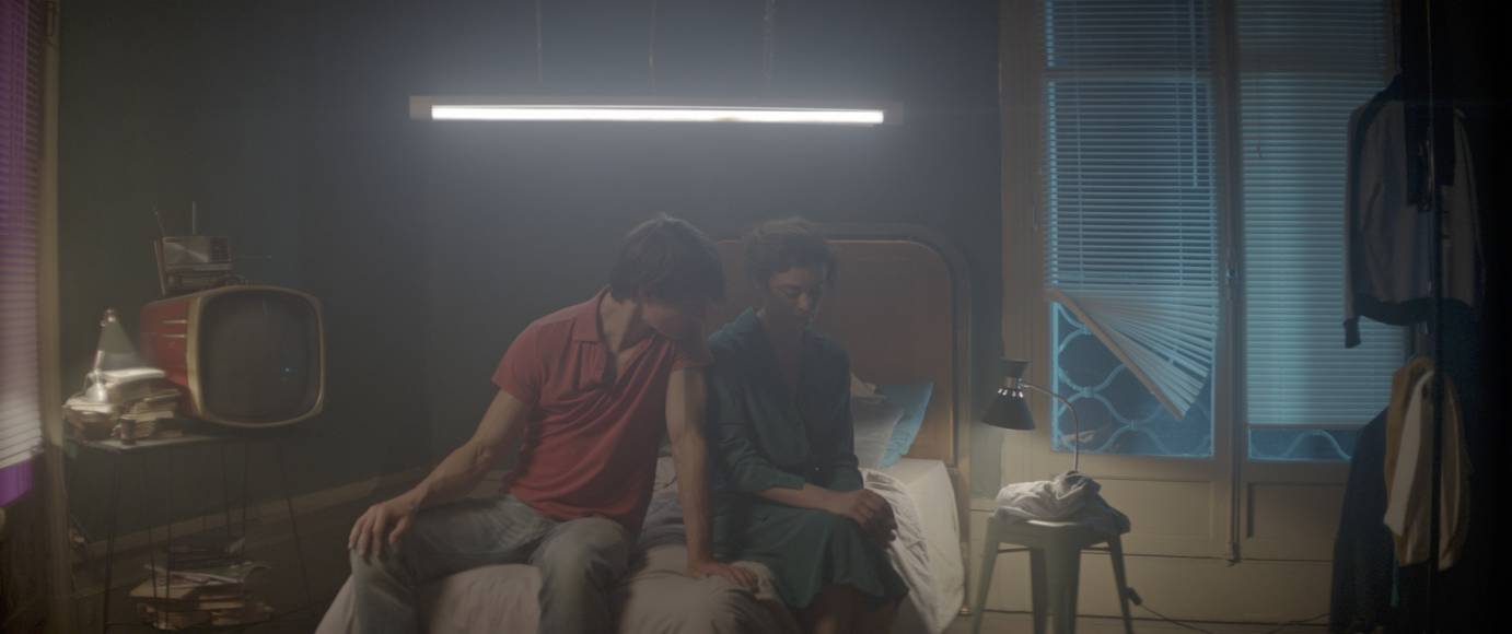 A man and a woman shrouded in fog sit on a bed as the man looks at the woman's shoulder