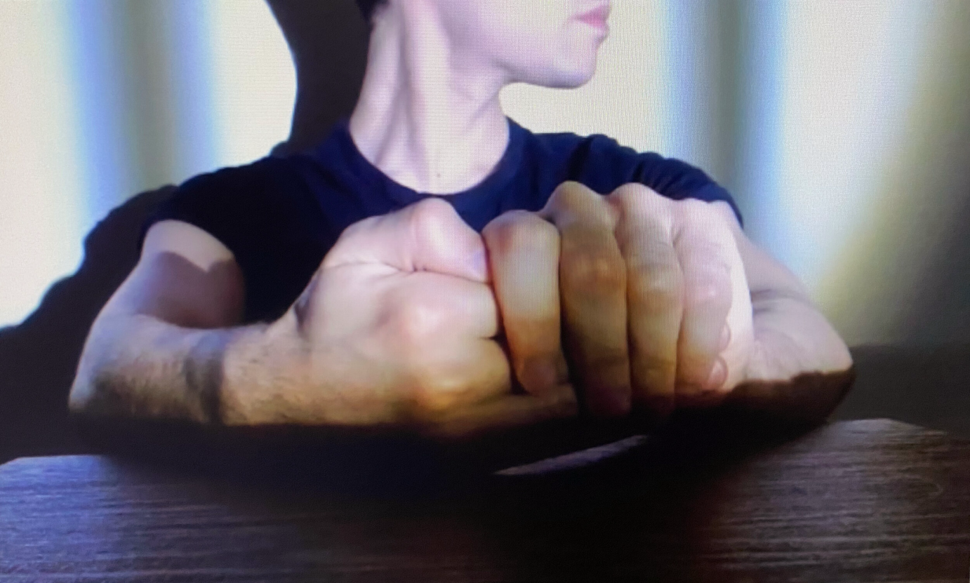 Part of a face shows against an illuminated background. Resting on a table, one hand clasps the thumb of the other