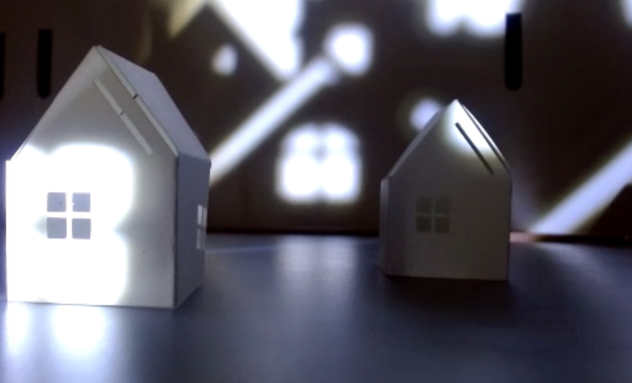 Two small white paper house with high roofs and a windows rest on a surface.