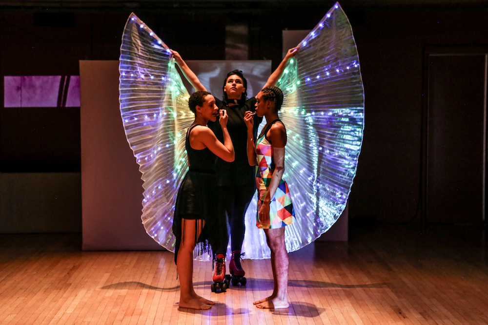 two Black women, one in a colorful mini dress, the other in a black outfit stare at one another as they put on their lipstick. Behind them is a figure on roller skates, wearing wings that are light up and make her seem as if she is an authortative angel