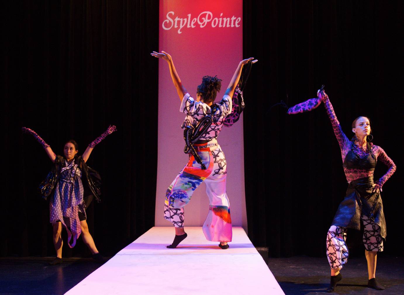 One woman stands on a runway, her back to the audience, and raises her arms in a V. There are two women on either side of her, off the runway and facing the audience.  